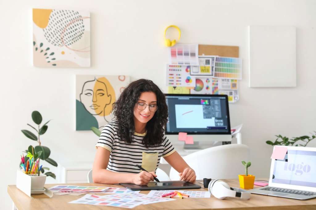 A woman designing at a home desk with a graphics tablet, desktop monitor, and mood board on the wall